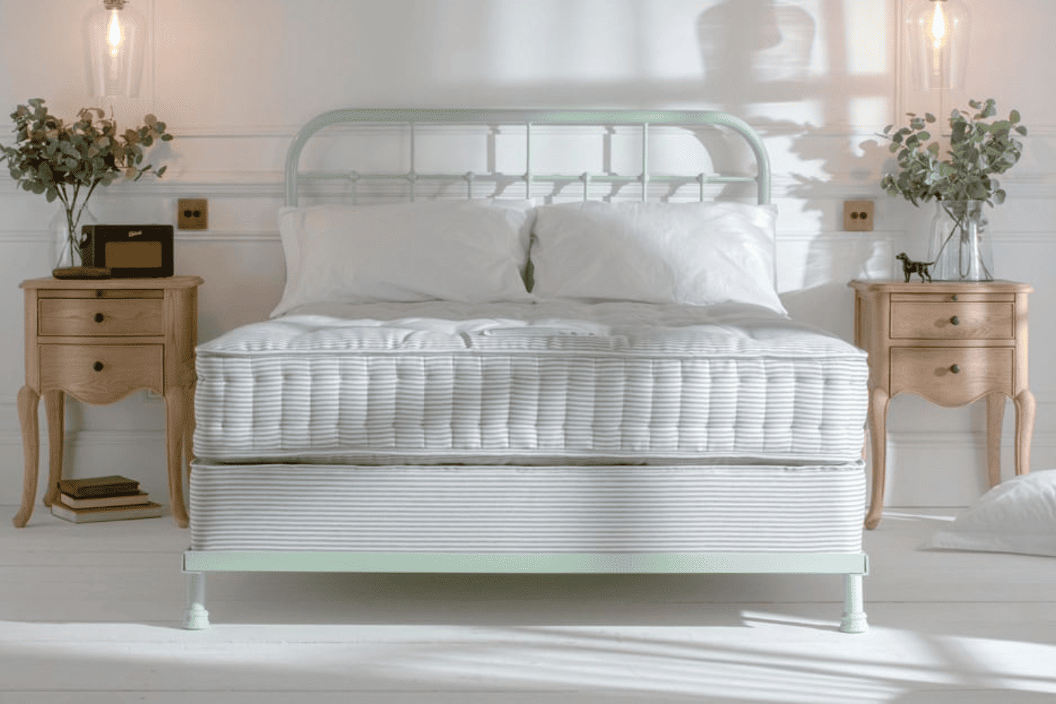 Mint green iron bed with mattress in a bright white bedroom, styled with wooden bedside tables and pendant lighting