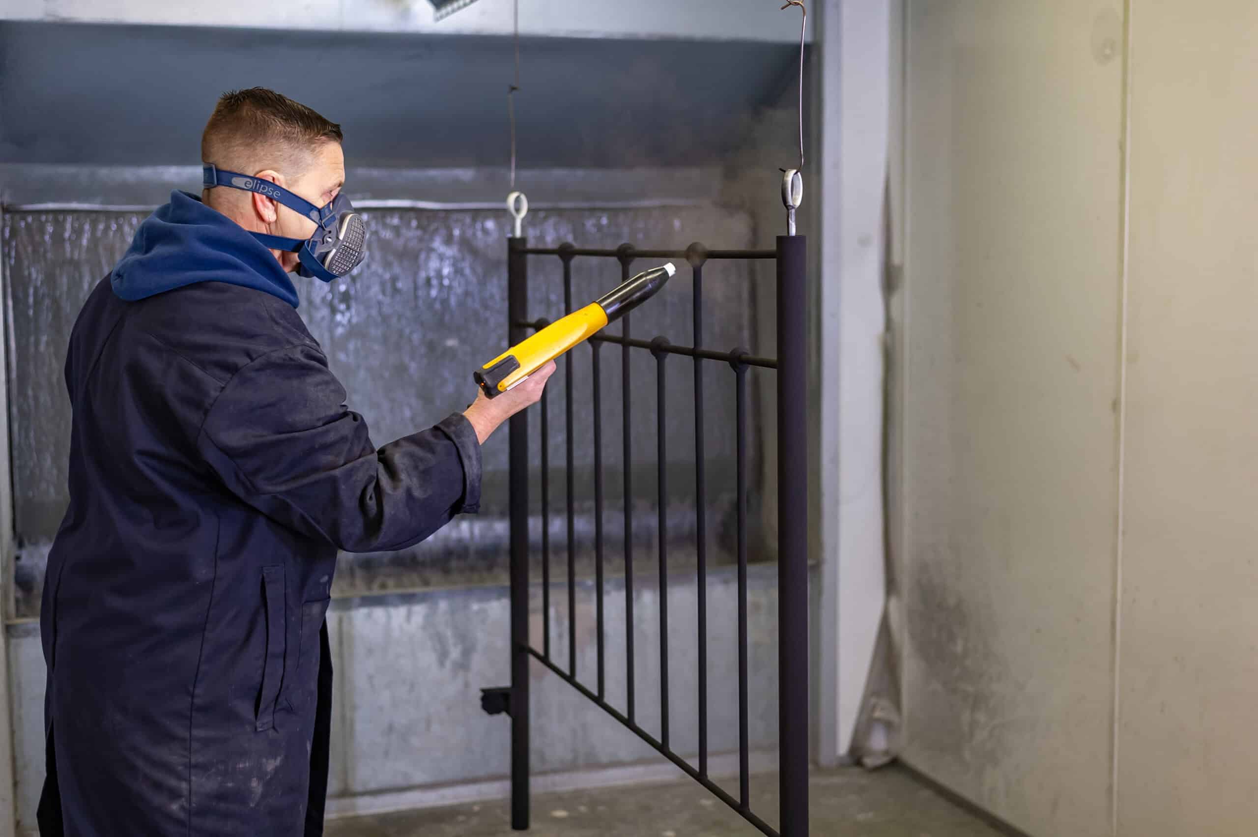 Skilled worker wearing safety gear inspecting a black iron bed frame with a measuring tool.